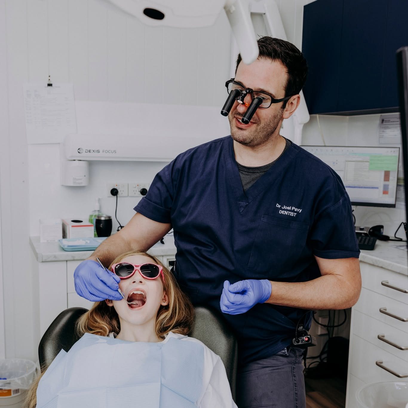 Dr Joel Pavy checking a young patient's teeth while she wears fun sunglasses